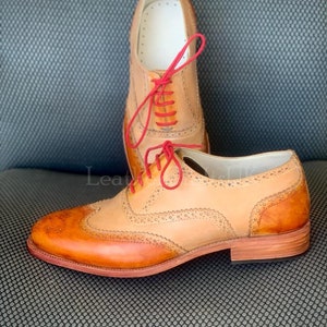 May include: A pair of tan leather Oxford shoes with red laces. The shoes have a wingtip design and a brogue pattern. The shoes are on a grey textured surface.