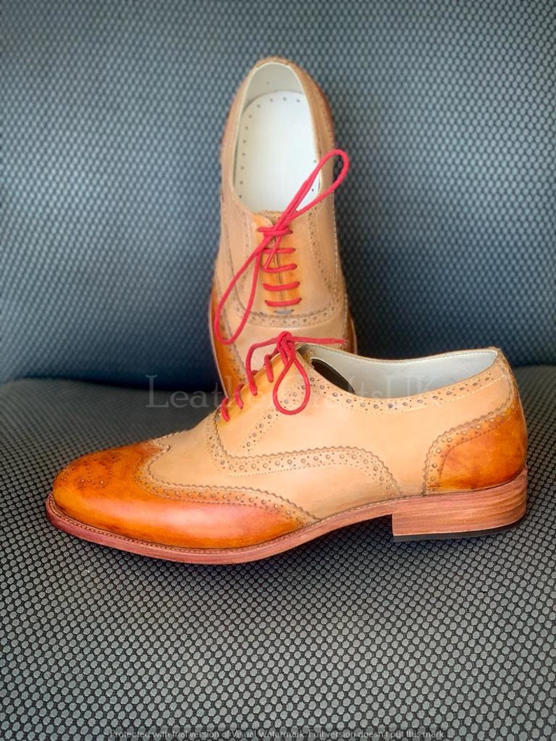 May include: A pair of tan leather Oxford shoes with red laces. The shoes have a wingtip design and a brogue pattern. The shoes are on a grey textured surface.