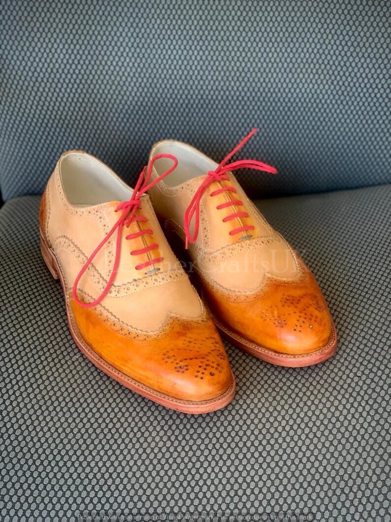 May include: A pair of tan leather brogue shoes with red laces. The shoes have a wingtip design and are made of a combination of tan and brown leather.