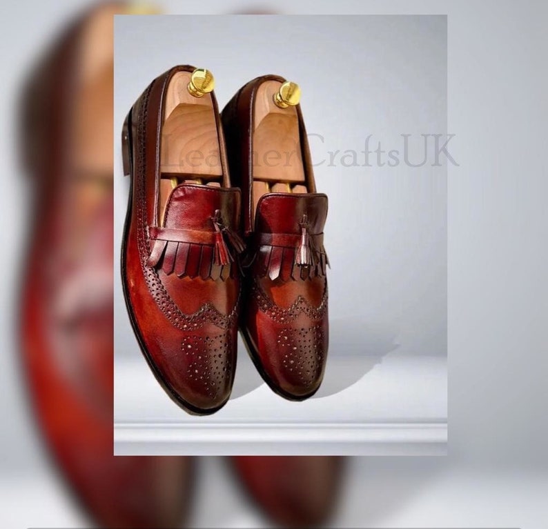 K&ouml;nnte beinhalten: Ein Paar burgunderfarbene Leder-Tasssel-Slipper mit Brogue-Details. Die Schuhe haben einen Fransen-Tasssel an der Vorderseite und sind aus hochwertigem Leder gefertigt.