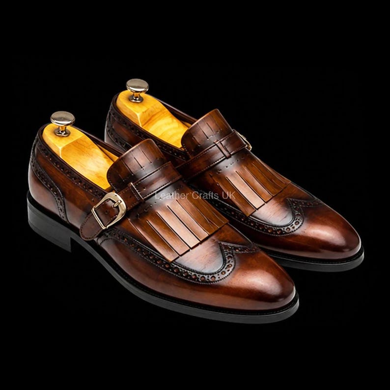 May include: A pair of brown leather monk strap shoes featuring a kiltie fringe detail, wingtip brogue perforations, a gold-toned metal buckle, and a wooden shoe tree insert. The shoes have a polished finish and a dark brown sole.