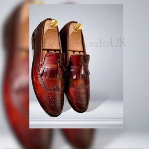 K&ouml;nnte beinhalten: Ein Paar burgunderfarbene Leder-Tasssel-Slipper mit Brogue-Details. Die Schuhe haben einen Fransen-Tasssel an der Vorderseite und sind aus hochwertigem Leder gefertigt.