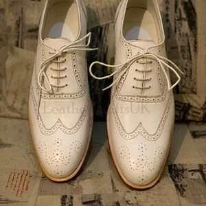 Puede incluir: Un par de zapatos de vestir de cuero blanco con cierre de cordones. Los zapatos tienen un dise&ntilde;o de punta de ala y est&aacute;n decorados con peque&ntilde;as perforaciones.