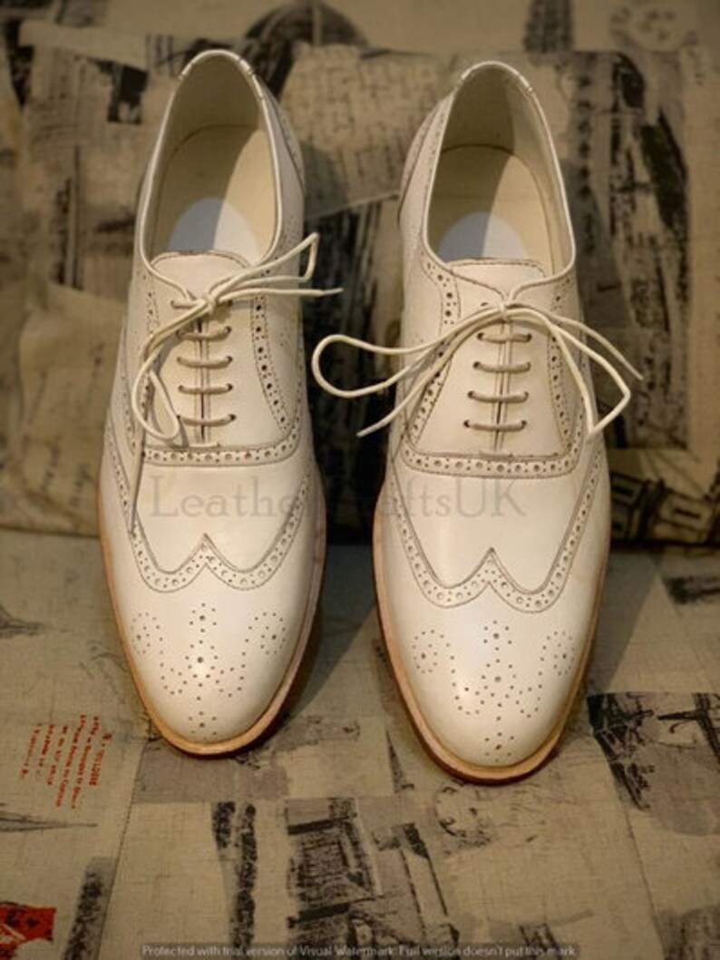 Puede incluir: Un par de zapatos de vestir de cuero blanco con cierre de cordones. Los zapatos tienen un dise&ntilde;o de punta de ala y est&aacute;n decorados con peque&ntilde;as perforaciones.