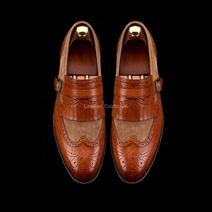 May include: A pair of tan leather kiltie loafers featuring wingtip brogue detailing, a decorative fringe panel, and a side buckle strap. The shoes have a smooth leather toe cap and a textured suede vamp, set against a dark background.