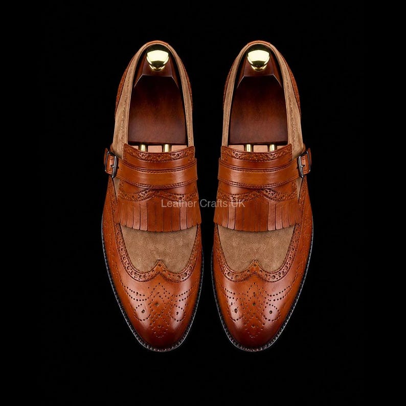 May include: A pair of tan leather kiltie loafers featuring wingtip brogue detailing, a decorative fringe panel, and a side buckle strap. The shoes have a smooth leather toe cap and a textured suede vamp, set against a dark background.