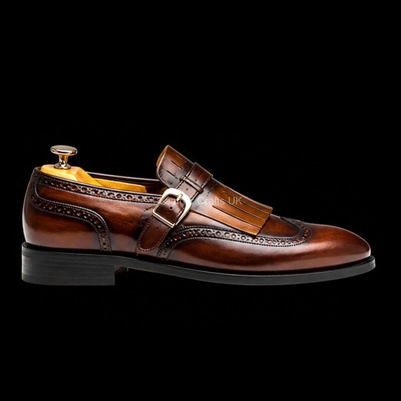 May include: A brown leather monk strap shoe featuring a kiltie fringe detail, a gold-toned metal buckle, and decorative brogue perforations along the wingtip toe cap and side panels. A wooden shoe tree is inserted into the heel area.