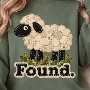 May include: An olive green crewneck sweatshirt featuring a central graphic of a sheep with a patchwork-style fleece made of cream and beige patterned circles. The sheep has a black head and legs, stands on green grass, and the word Found. in black.