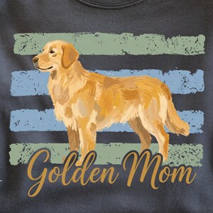 May include: A charcoal gray t-shirt featuring a golden retriever illustration centered over horizontal distressed stripes in sage green and light blue. The text "Golden Mom" is printed in a cursive gold font below the dog graphic.
