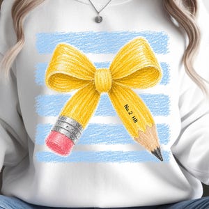 Puede incluir: Una sudadera blanca de cuello redondo con un gráfico de un lápiz amarillo atado en forma de lazo. El lápiz tiene una virola plateada, una goma rosa y una punta afilada con el texto No. 2 HB impreso, sobre un fondo de rayas azul claro.