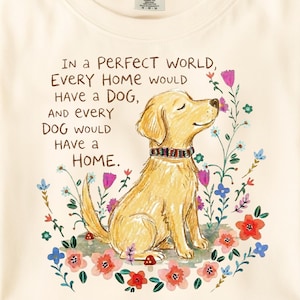May include: A cream-colored t-shirt featuring a golden dog illustration and the text: In a PERFECT WORLD, EVERY HOME WOULD HAVE A DOG, AND EVERY DOG WOULD HAVE A HOME. The design includes colorful hand-drawn flowers surrounding the dog.