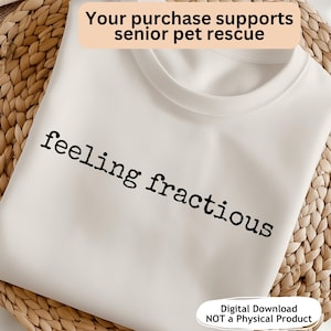 Può includere: T-shirt color crema con la scritta nera "feeling fractious". La maglietta ha uno scollo tondo ed è esposta su una superficie tessuta. Il testo in alto recita "Your purchase supports senior pet rescue". Il testo in basso recita "Digital Download NOT a Physical Product".