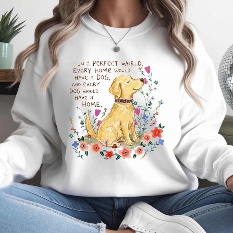 May include: A white crewneck sweatshirt featuring a graphic illustration of a golden dog sitting among colorful flowers. The text reads: In a PERFECT WORLD, EVERY HOME would HAVE a DOG, AND EVERY DOG WOULD HAVE a HOME. The dog wears a patterned collar.