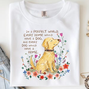 May include: A white t-shirt featuring a graphic of a golden dog wearing a colorful collar, surrounded by various flowers. The text reads: IN A PERFECT WORLD, EVERY HOME WOULD HAVE A DOG, AND EVERY DOG WOULD HAVE A HOME. The shirt has a crew neckline.