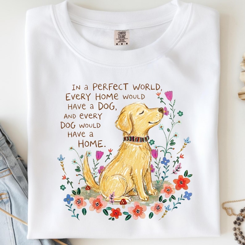 May include: A white t-shirt featuring a graphic of a golden dog wearing a colorful collar, surrounded by various flowers. The text reads: IN A PERFECT WORLD, EVERY HOME WOULD HAVE A DOG, AND EVERY DOG WOULD HAVE A HOME. The shirt has a crew neckline.
