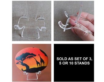 May include: Clear acrylic display stands, perfect for showcasing small items. The stands are shown holding a painted rock with a sunset and giraffe design. Text on a black background reads "SOLD AS SET OF 3, 5 OR 10 STANDS."