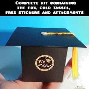 May include: A black graduation cap-shaped box kit featuring a gold tassel and a circular sticker with the text We did it! and a small graduation cap icon. The kit includes the box, gold tassel, stickers, and attachments for assembly.