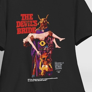Puede incluir: Camiseta negra con un diseño de póster de película vintage con un título rojo "THE DEVIL'S BRIDE" y un gráfico de una mujer que es llevada por una figura con cuernos.