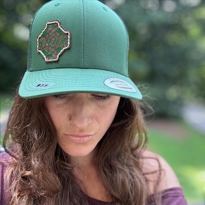 May include: A solid green baseball cap featuring a brown leather patch with an embossed Celtic cross design. The cap has a structured front panel, a flat brim, and a circular sticker on the brim. The design includes intricate knotwork patterns.