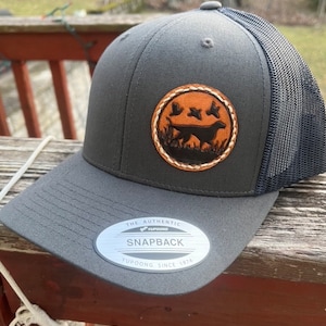 May include: A charcoal gray snapback cap with a mesh back and a circular leather patch featuring a silhouette of a hunting dog and three flying birds. The brim displays a silver sticker with the text THE AUTHENTIC YUPOONG SNAPBACK YUPOONG SINCE 1974.