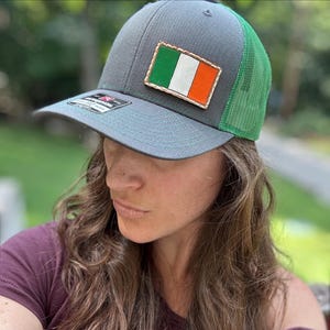 May include: A gray baseball cap with a green mesh back and a rectangular patch featuring the green, white, and orange Irish flag. The cap has a flat brim with a small black sticker on the left side and a structured front panel with stitching.