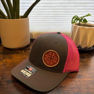 May include: A charcoal gray trucker hat with a bright pink mesh back and matching pink stitching on the brim. The front features a circular brown leather patch embossed with an intricate Celtic knot design and a Richardson 112 sticker on the brim.