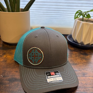 May include: A charcoal gray trucker hat with a bright turquoise mesh back and matching turquoise stitching on the brim. The front features a circular patch with a brown leather border and a turquoise Celtic knot design. A Richardson 112 label is attached.