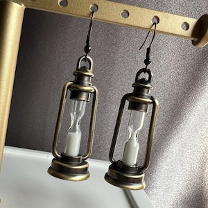 May include: A pair of gold-toned lantern-shaped earrings with a small hourglass inside each lantern. The earrings are hanging from a display stand.