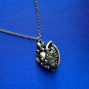 May include: A silver necklace with a steampunk-style heart pendant. The pendant is detailed with gears, pipes, and other mechanical elements.