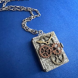 May include: A silver-toned steampunk pendant necklace. The rectangular pendant features a clock face and gears in bronze and copper tones. The pendant hangs from a silver chain against a blue background. The pendant has an ornate, engraved design.