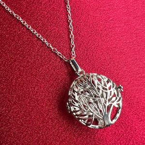 May include: A silver-colored pendant necklace featuring a detailed tree of life design within a circular frame. The pendant hangs from a delicate chain. The background is a solid red color.