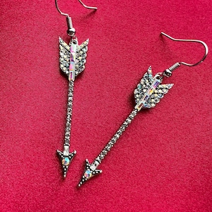 May include: A pair of silver-toned arrow-shaped dangle earrings featuring clear rhinestones along the shaft and fletching. The arrowheads and fletching are accented with iridescent, rectangular-cut crystals and small round stones.