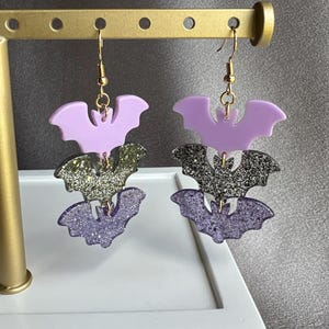Può includere: Un paio di orecchini pendenti con tre ciondoli a forma di pipistrello sovrapposti. Il pipistrello superiore è di colore viola chiaro, quello centrale è glitterato argento e quello inferiore è glitterato viola. Uniti da anellini in metallo dorato.
