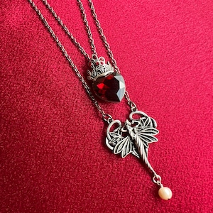 May include: A silver-toned necklace featuring a faceted dark red heart-shaped pendant topped with a crown-like setting, connected to a silver-toned fairy figure with wings and a small dangling white pearl bead at the bottom of the pendant.