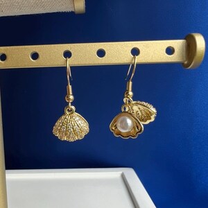 May include: A pair of gold-toned drop earrings featuring a seashell design. One earring is a closed shell encrusted with small clear crystals, while the other is an open shell revealing a single white faux pearl. Both hang from gold-toned hooks.
