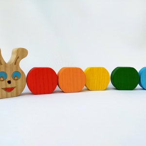 May include: A wooden toy caterpillar with a smiling face and blue eyes. The caterpillar is made up of six colorful segments: red, orange, yellow, green, blue, and brown.