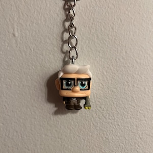 May include: A white and brown figurine keychain of an older man with glasses and a robotic arm. The keychain is attached to a silver chain and ring.