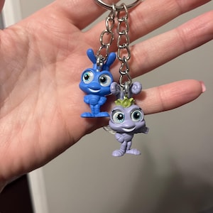 May include: A keychain featuring two small character figurines. One is blue, the other is purple. Both have large eyes and are attached to a silver chain and keyring. The figurines are likely from a children's animated movie.