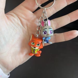 May include: A keychain featuring two cartoon character figurines: a fox in orange and a rabbit in gray and blue. The figures are attached to a silver metal chain and keyring. The fox has a green scarf, and the rabbit has purple eyes.
