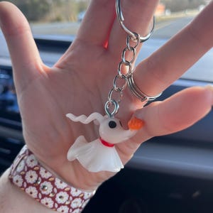 May include: A white Zero keychain from The Nightmare Before Christmas. The keychain features a small, translucent Zero figure with a red collar and an orange nose. The keychain is attached to a silver metal ring and chain.