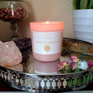 Puede incluir: Una vela rosa con una etiqueta blanca que dice "The Many Phases Queen of Pentacles Cedarwood and Vanilla". La vela está en una bandeja plateada con pétalos de rosa y un cristal rosa.