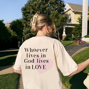 Könnte beinhalten: Ein cremefarbenes Kurzarm-T-Shirt mit schwarzem Text auf der Rückseite, der "Whoever lives in God lives in LOVE" lautet und über einem hellrosa Herzmotiv liegt. Das T-Shirt hat einen Rundhalsausschnitt und eine lockere, entspannte Passform.
