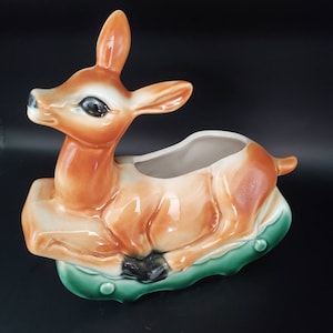 May include: A ceramic planter shaped like a brown deer lying down on a green base. The deer has black eyes and a white belly. The planter has a hole in the top for planting.