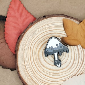May include: A mushroom-shaped enamel pin with a white cap and grey details, set on a wooden slice with decorative paper leaves. The pin features a detailed design with a dark grey stem and cap accents.