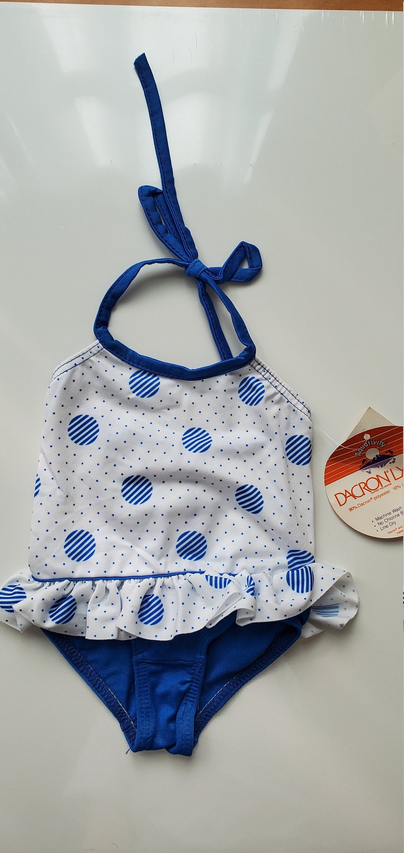 May include: A blue and white polka dot swimsuit with a ruffled bottom. The swimsuit has a halter top with a blue tie. The tag says "Dacron Ly 90% Dacron polyester 10% Machine Wash No Chlorine Bleach Line Dry."