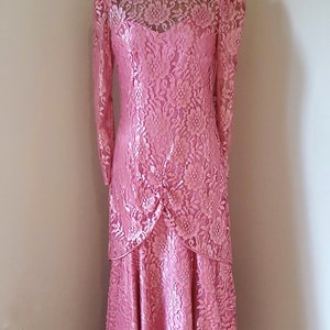 May include: A pink lace dress with a high neckline and long sleeves. The dress has a tiered skirt and is displayed on a mannequin.