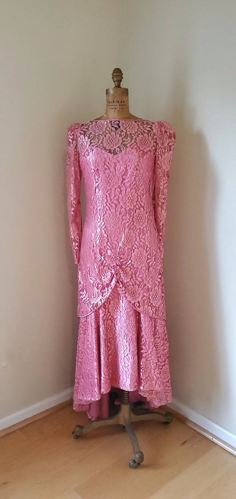 May include: A pink lace dress with a high neckline and long sleeves. The dress has a tiered skirt and is displayed on a mannequin.