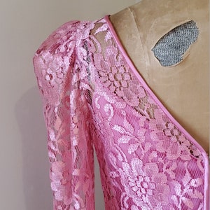 May include: A pink lace dress with a V-neckline and long sleeves. The dress has a delicate floral pattern and a fitted silhouette.