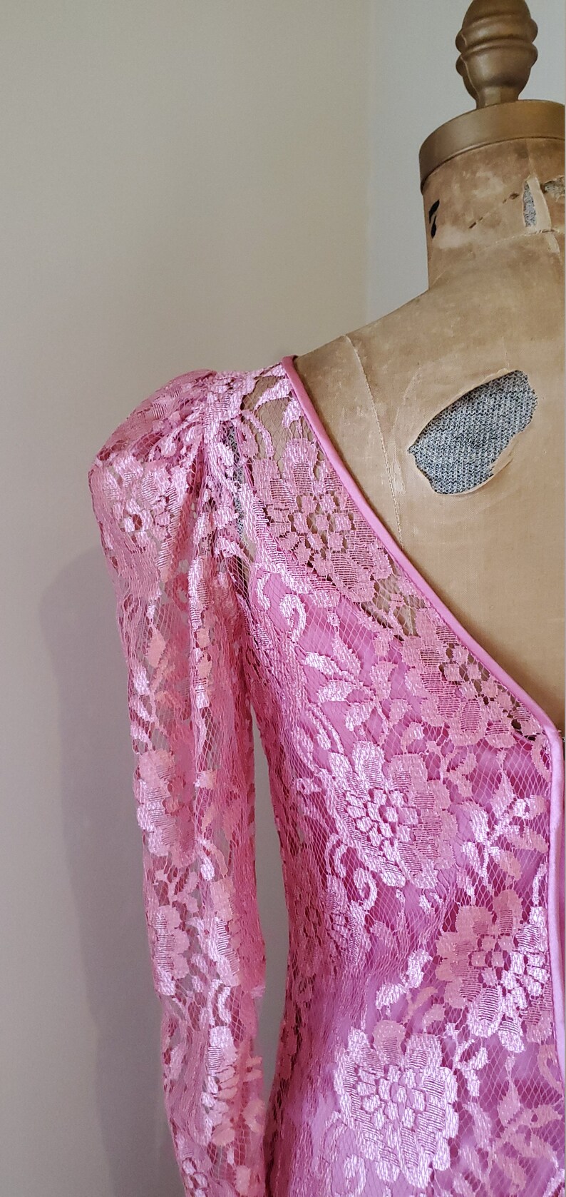 May include: A pink lace dress with a V-neckline and long sleeves. The dress has a delicate floral pattern and a fitted silhouette.