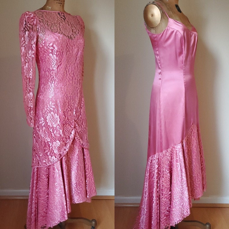 May include: A pink satin and lace dress with a long sleeve and a high neckline. The dress has a fitted bodice and a flowing skirt with a high-low hemline.